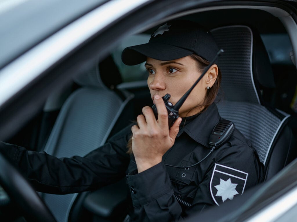 Policewoman using a radio while patrolling in a security vehicle, representing mobile patrol services by International Security Services in North Texas.