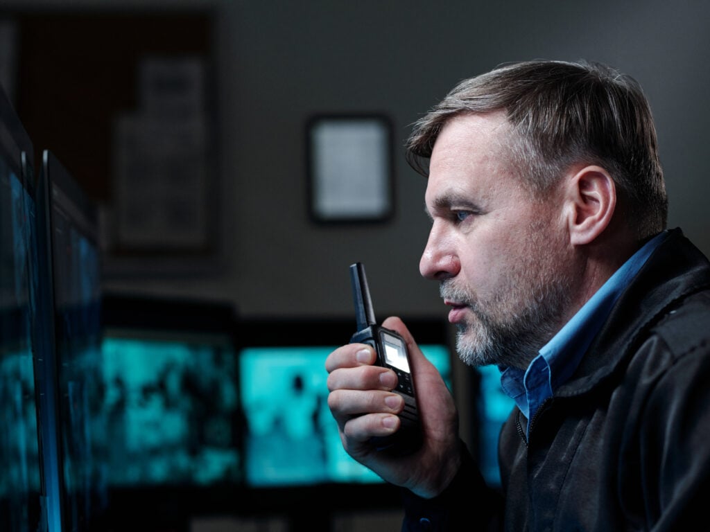 Security guard communicating via walkie-talkie, monitoring surveillance screens in a control room, emphasizing professional security services.