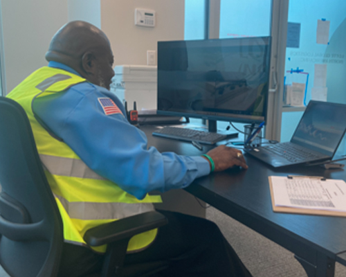 Security officer monitoring community activity at a desk with a computer, wearing a reflective vest, emphasizing community safety and communication for International Security Services.